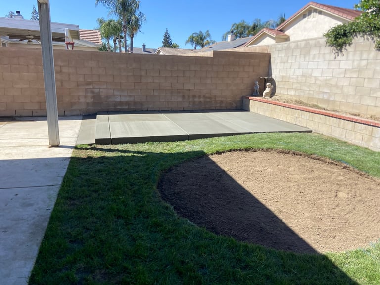 Backyard under renovation with new sod, mulch bed, concrete patio, and brick walls under clear blue sky