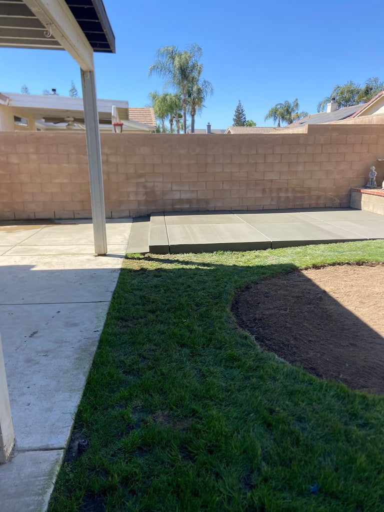 Backyard patio with concrete pad, pergola, brick wall, newly landscaped lawn, and palm trees under clear blue sky
