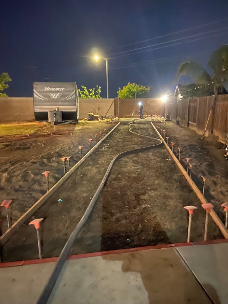 Nighttime construction site with surveying equipment, irrigation lines, and wooden stakes on sandy ground with parked trailer and street lighting