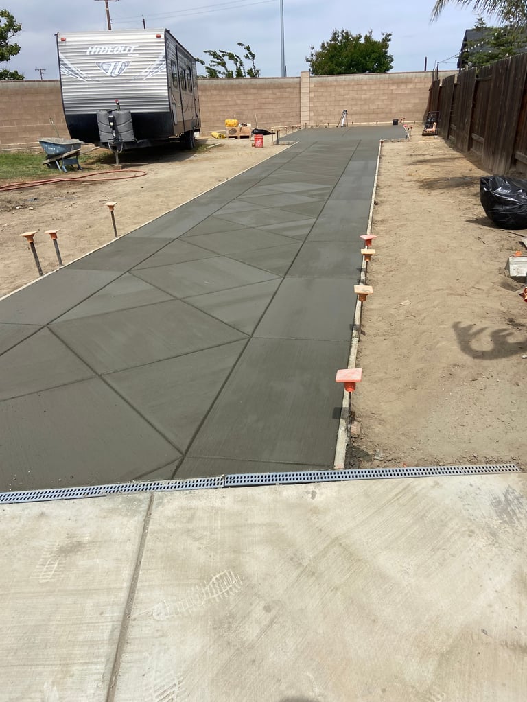 Concrete driveway being poured in a residential backyard with construction trailer, wooden fencing, and dirt ground visible