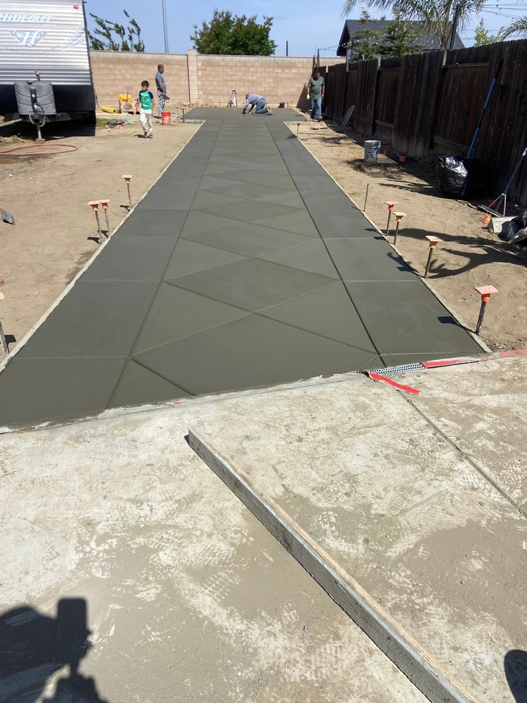 Fresh concrete being poured for a long driveway in a residential area with wooden fences and houses visible on both sides